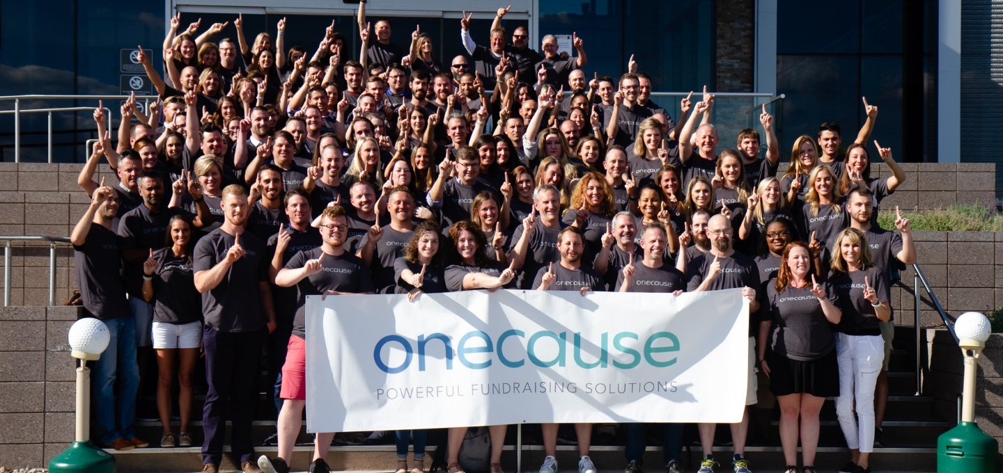 A large group of people stand on an outdoor staircase holding a sign with the OneCause logo.