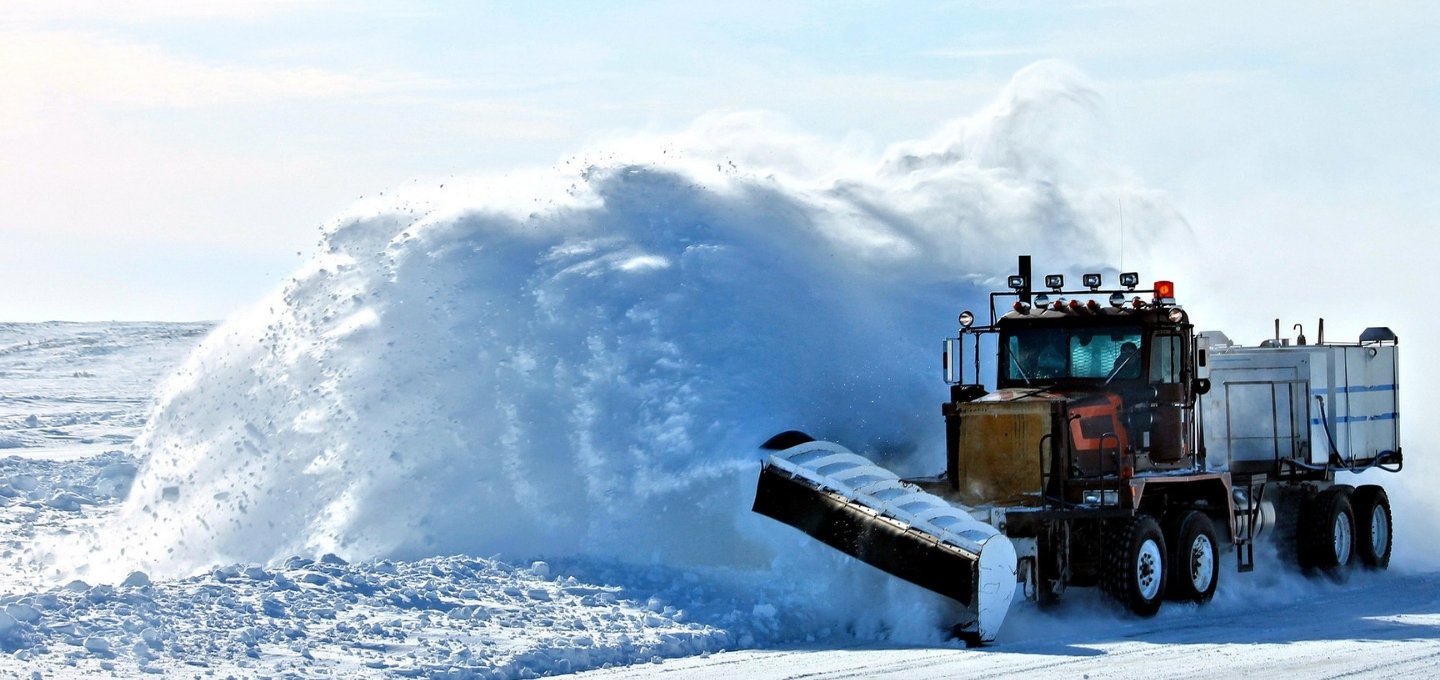A large snow plow is digging into snow on the side of a country road, sending up a plume of white snow.