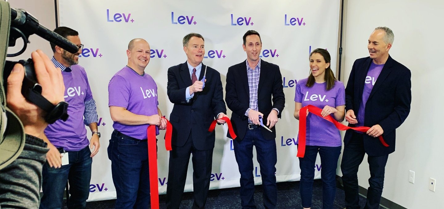 A group of people stand in front of the logo for Lev while cutting a large ribbon.