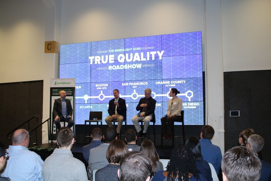 A panel of speakers sits on a stage with True Quality Roadshow displayed behind them.