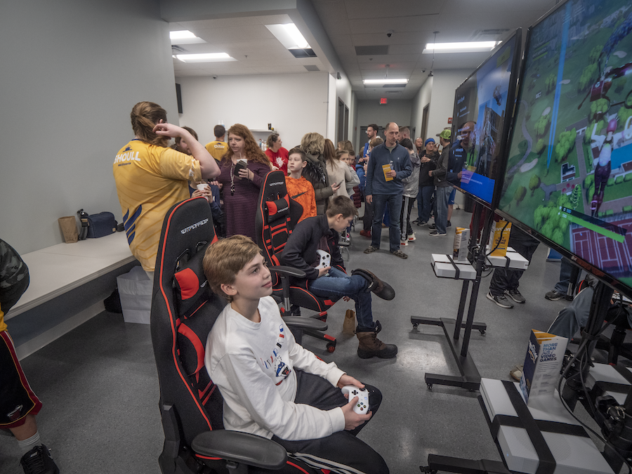 A group of young children sit in gaming chairs and play games on TVs while their parents watch.