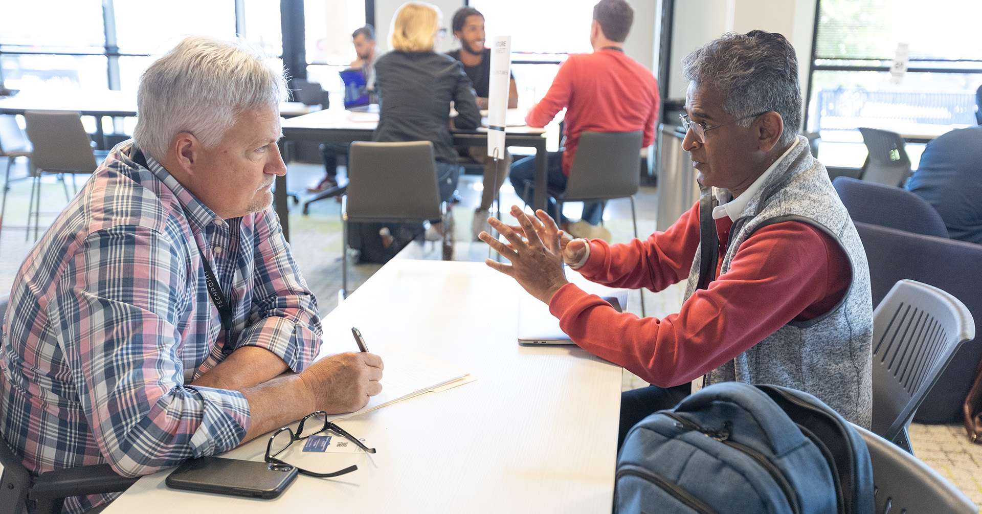 Two men are seated across from each other at a table in a bright, modern meeting space, engaged in a focused conversation. One man gestures expressively while the other listens and takes notes. Several small group discussions are happening in the background.
