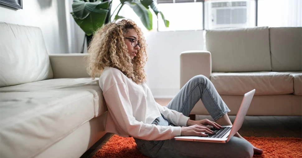 A young woman sits on an orange rug in a bright living room, leaning against a cream-colored couch while typing on a laptop. Sunlight comes through large windows behind her, and green houseplants are visible in the background.