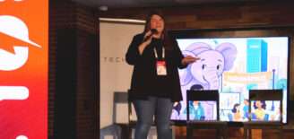 Sally Reasoner speaks on stage at the BOLT Conference, powered by gener8tor, hosted by TechPoint. She stands with a microphone and event badge in front of a large screen featuring an illustrated Indianapolis cityscape with a purple elephant and “Indianapolis” banner. A red BOLT-branded display and TechPoint backdrop are visible behind her