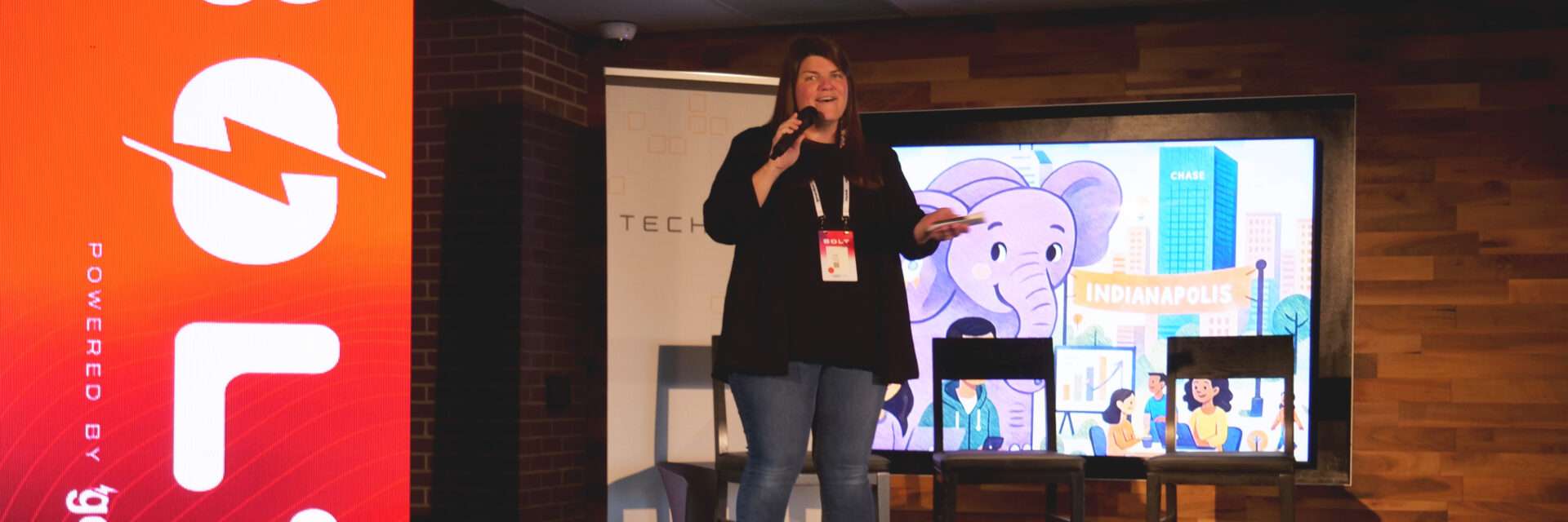 Sally Reasoner speaks on stage at the BOLT Conference, powered by gener8tor, hosted by TechPoint. She stands with a microphone and event badge in front of a large screen featuring an illustrated Indianapolis cityscape with a purple elephant and “Indianapolis” banner. A red BOLT-branded display and TechPoint backdrop are visible behind her