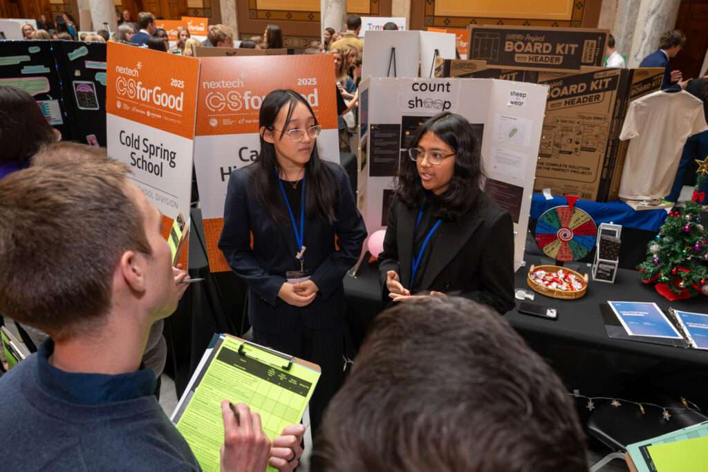 Two high school students present their CSforGood project to event judges, standing beside display boards and signage for the High School Division inside the Indiana Statehouse.