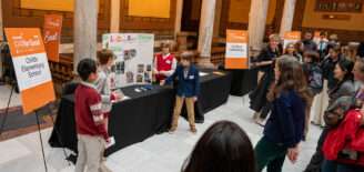 Elementary students present their CSforGood project to a crowd inside the Indiana Statehouse, standing beside display boards and event signage for the Nextech competition.