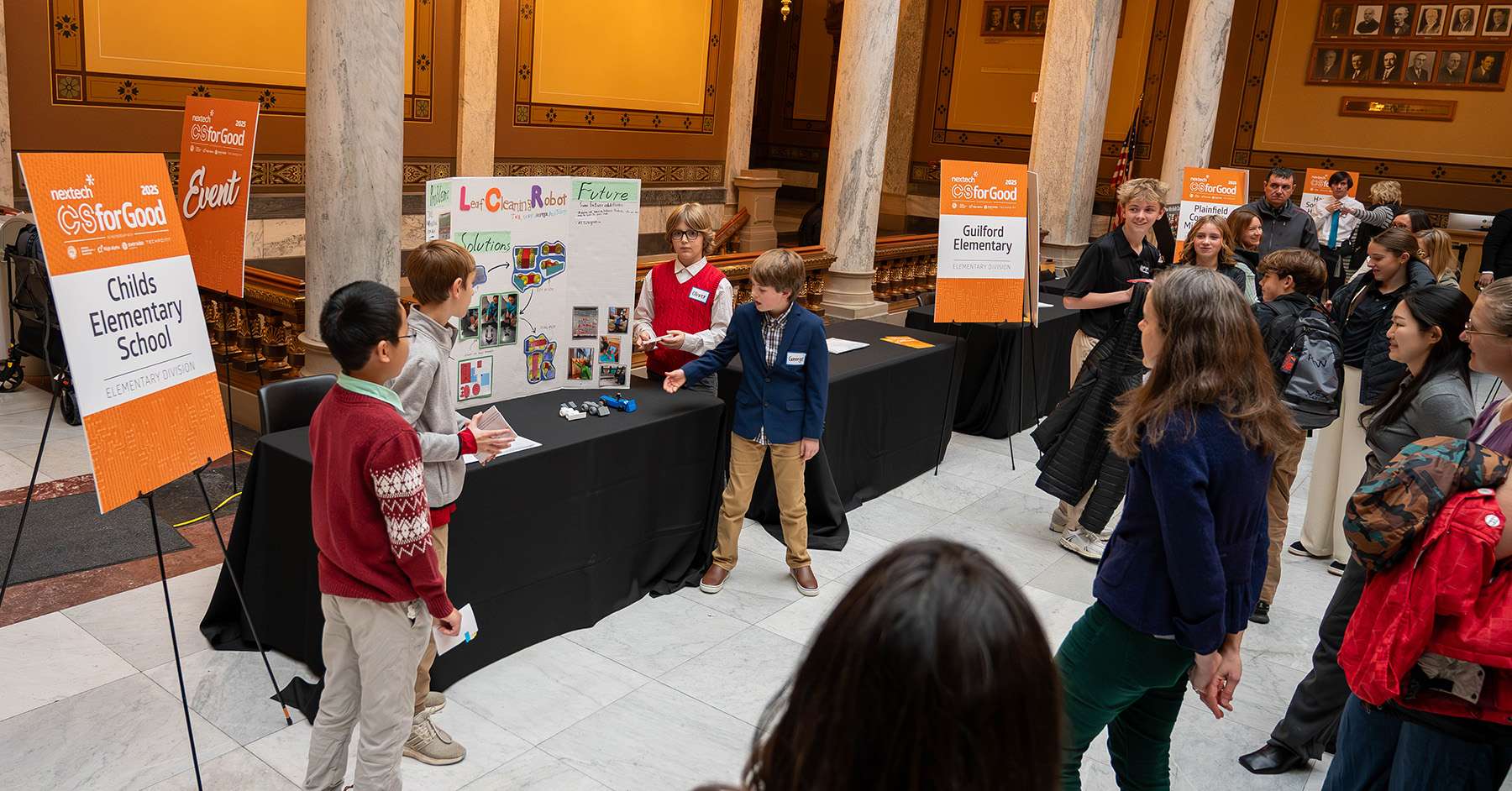 Elementary students present their CSforGood project to a crowd inside the Indiana Statehouse, standing beside display boards and event signage for the Nextech competition.