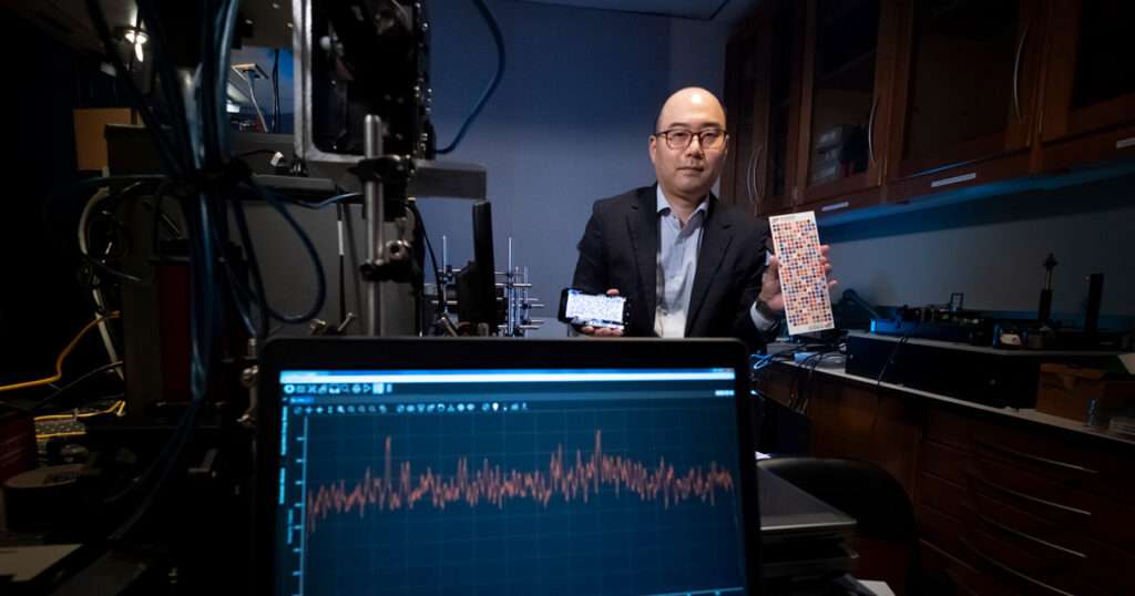 A Purdue University engineer sits in a laboratory holding a smartphone and a patterned diagnostic test strip, with lab instruments and a data graph on a monitor in the foreground, illustrating work on a new technology aimed at earlier identification of preeclampsia risk.