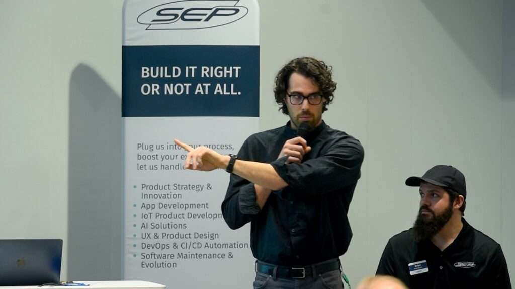 Two SEP team members present a case study at an event, with one speaker pointing toward the audience while holding a microphone and another seated beside him, in front of a SEP banner.