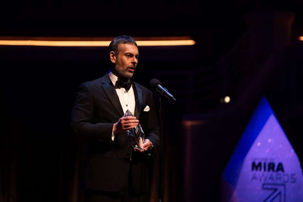 Raman Ohri stands on stage in formal attire, speaking at a microphone while holding the Exceptional Employer of the Year Mira Award during the 2025 ceremony.