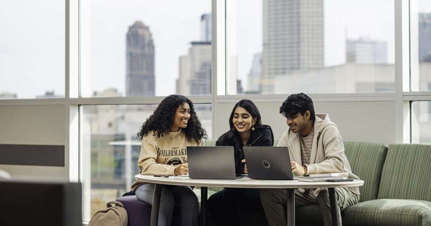 Three Purdue students collaborating on laptops in a modern campus space with the Indianapolis skyline visible through large windows.