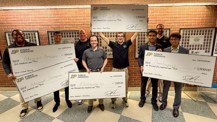 Group photo of Purdue Innovates student teams holding oversized award checks (including Plug N’ Play, AeroInspect, and Electrocean Inc.) in a hallway with a brick wall and Purdue University photo displays.