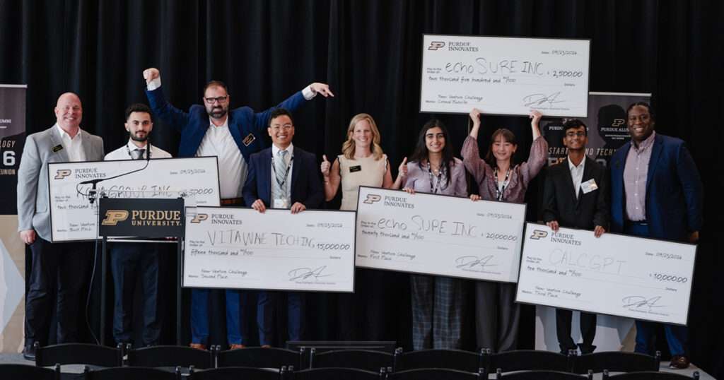 Eight people face the camera as students hold five oversized checks.