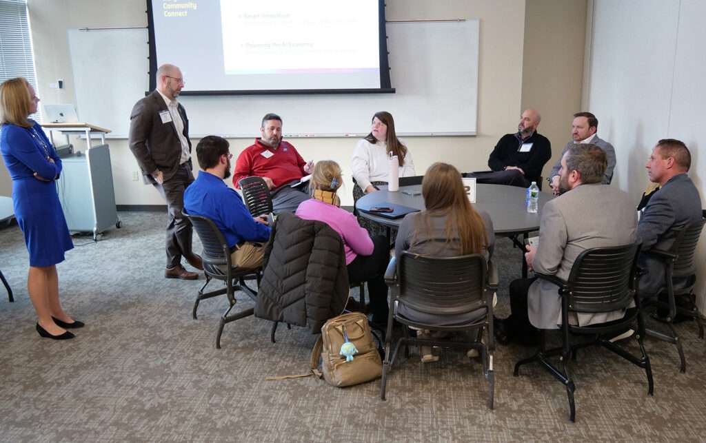 TechPoint Community Connect attendees participate in a breakout session, gathered around a table in small-group discussion while a presenter stands nearby.
