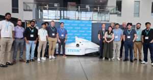 Sallie Mae team members and their Xterns pose in front of a white Xtern-branded IndyCar and blue Xtern backdrop at Dallara.