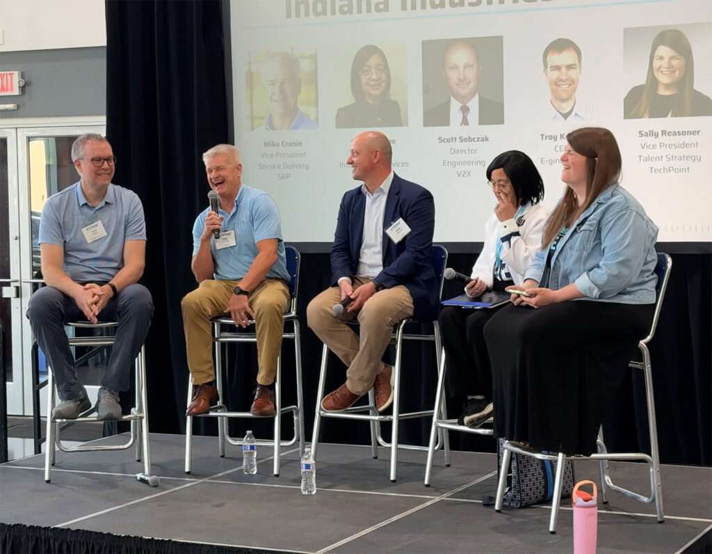 Five tech leaders sit together on a stage speaking together as panelists.