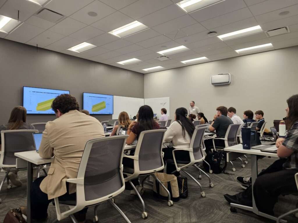 Participants at laptops watch a speaker present a 3D CAD model on dual screens during a technical workshop session.