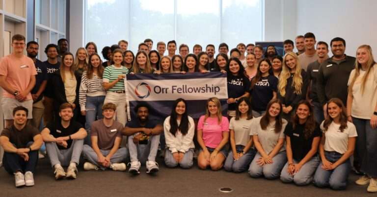 A large group of Orr fellows gather indoors, holding an Orr Fellowship banner.