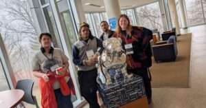 Four people wearing CCOM conference badges pose with a silver Butler bulldog statue inscribed with communication-related fields.