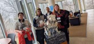 Four people wearing CCOM conference badges pose with a silver Butler bulldog statue inscribed with communication-related fields.