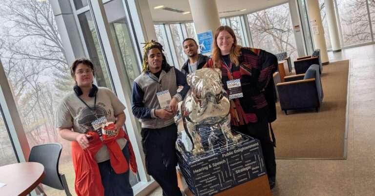 Four people wearing CCOM conference badges pose with a silver Butler bulldog statue inscribed with communication-related fields.