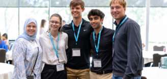 Five attendees stand together and smile for a photo at an XTERN event. Three wear blue XTERN lanyards and name badges, with event tables and large windows visible in the background.