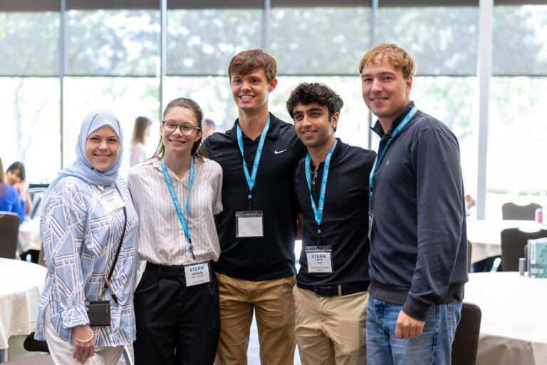Five attendees stand together and smile for a photo at an XTERN event. Three wear blue XTERN lanyards and name badges, with event tables and large windows visible in the background.