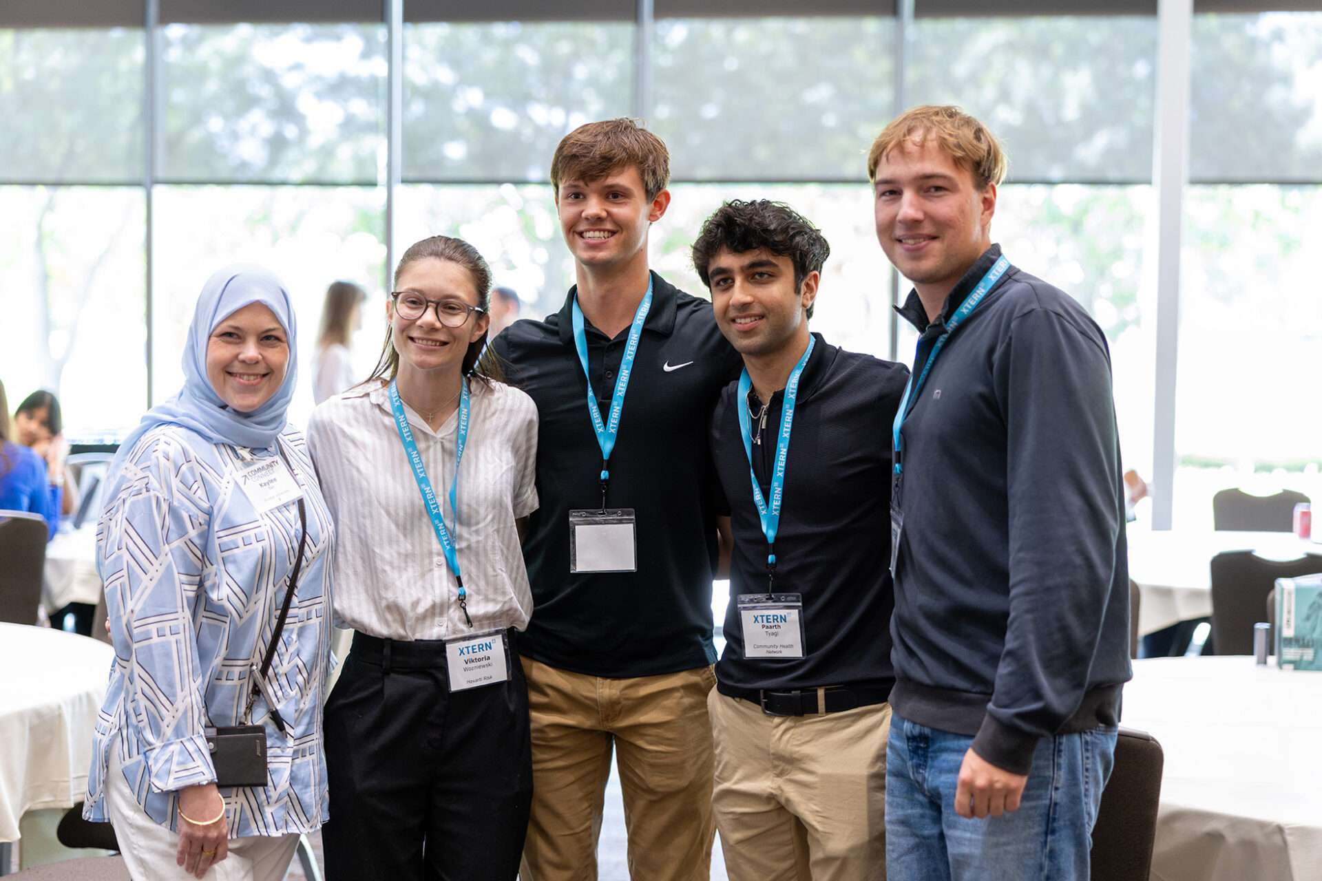 Five attendees stand together and smile for a photo at an XTERN event. Three wear blue XTERN lanyards and name badges, with event tables and large windows visible in the background.