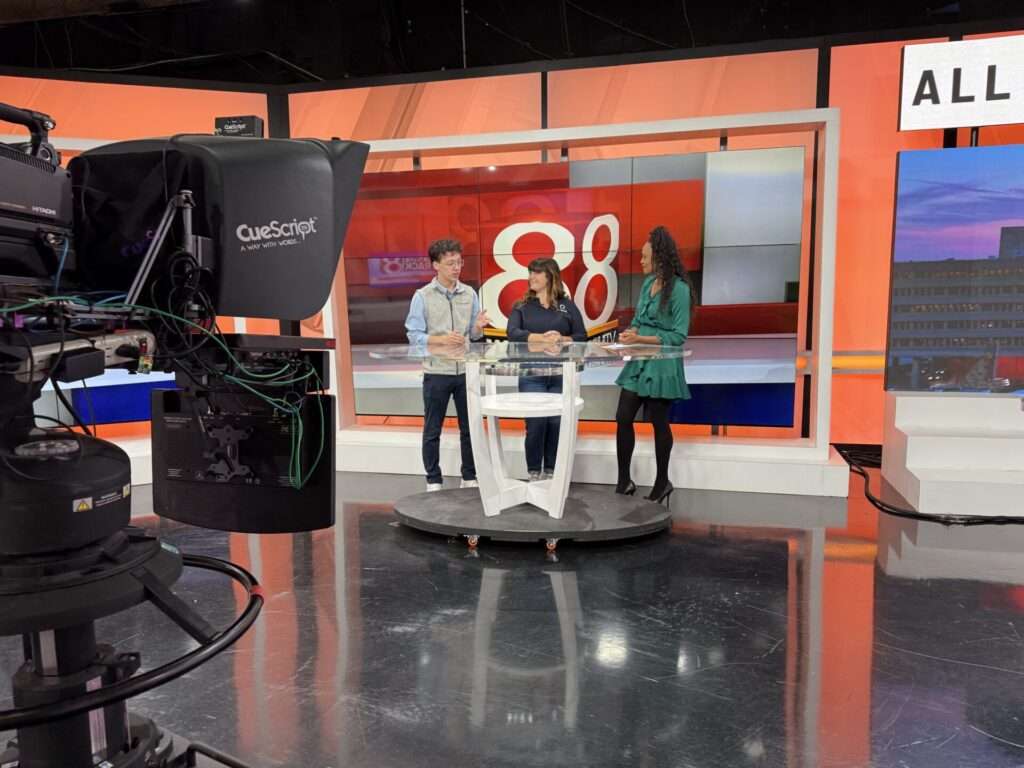 Three people stand at a news desk during a segment on a local television station, with on-air graphics visible in the background.