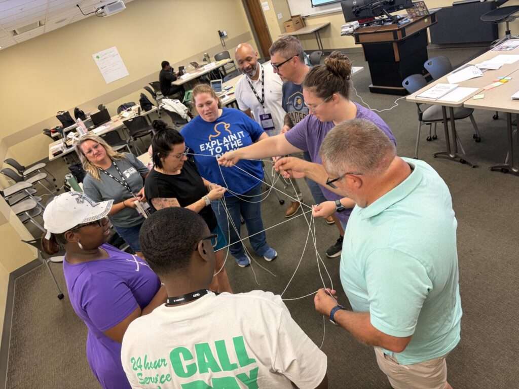 A group of educators works together to untangle a web of strings during a hands-on team-building exercise in a classroom.

