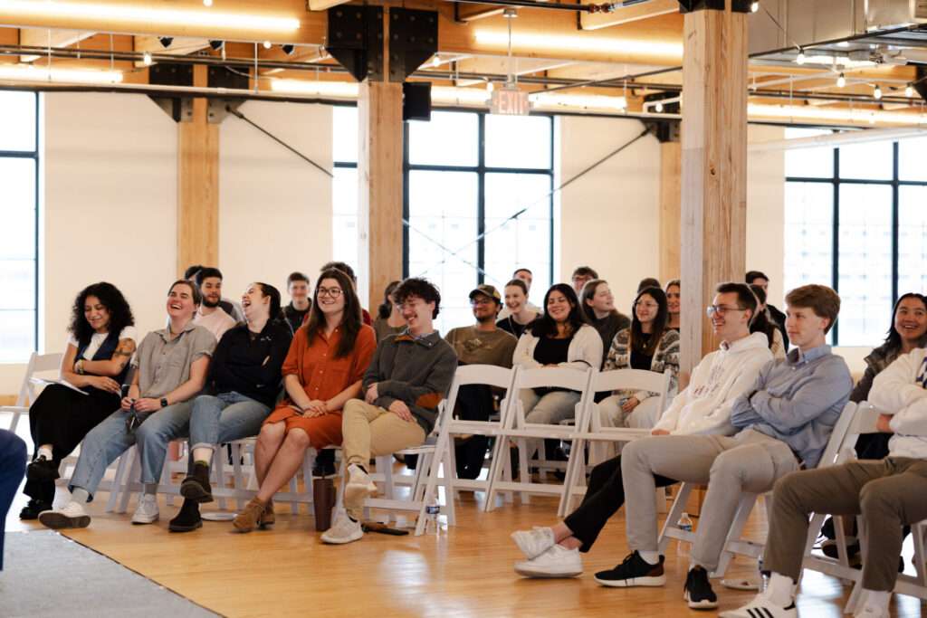 A group of attendees sit and laugh together during a presentation in an open, industrial-style event space. 