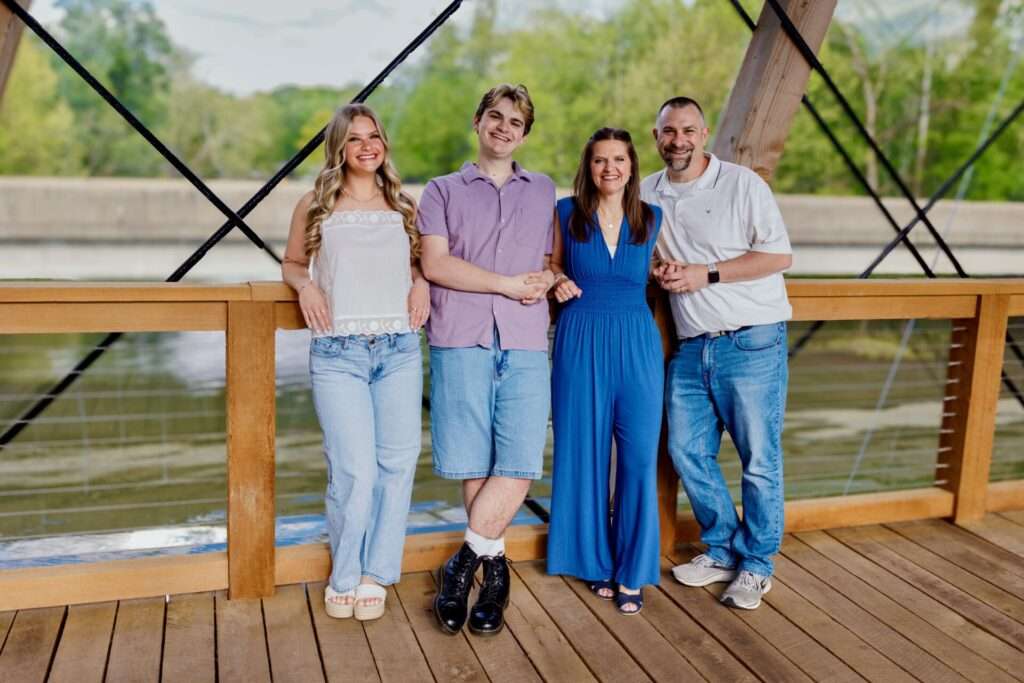 Nick Fishel poses with his wife and two children on a wooden pedestrian bridge, smiling together outdoors.