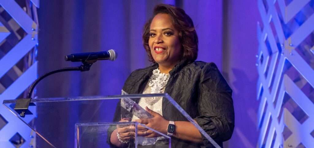 Akilah Darden, founder of The Darden Group LLC, smiles while holding a Mira Award trophy at a podium.