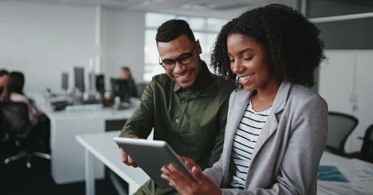 Two coworkers review information together on a tablet in a modern office.