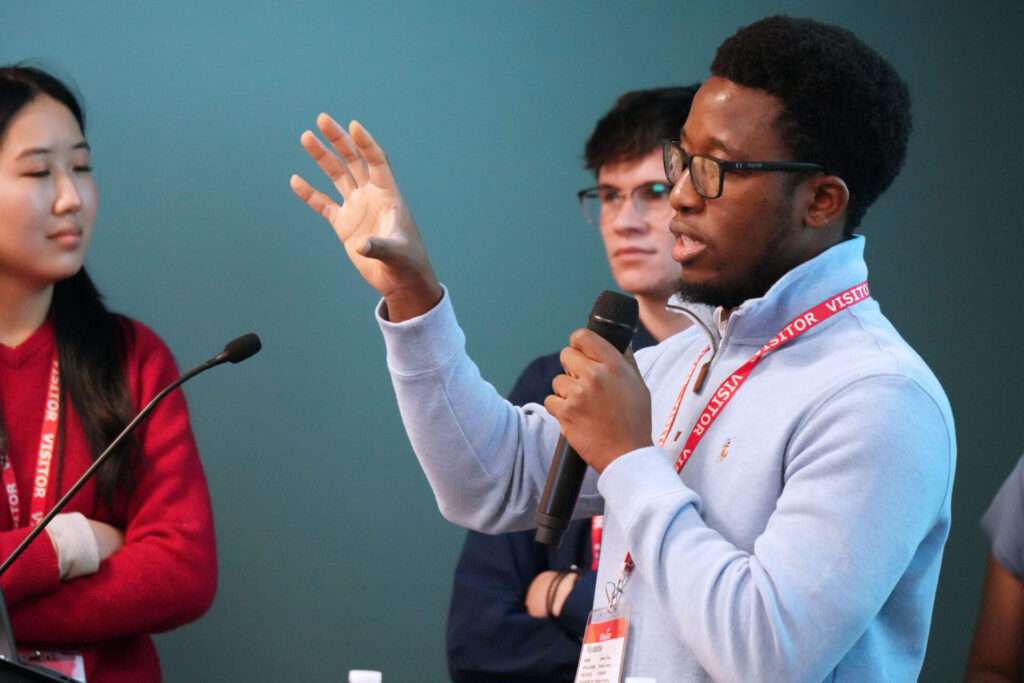 A student speaks into a handheld microphone while gesturing during a group discussion at the MISO Xtern Challenge. Other students listen nearby.