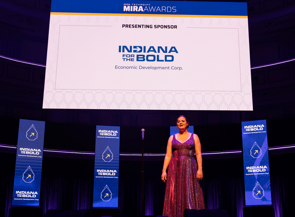 Chelsea Linder stands onstage in a sparkling gown at the 2026 Mira Awards, recognizing presenting sponsor Indiana Economic Development Corporation, whose logo appears on screens behind her.